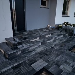 DB.Building Dominik Budzyński Usługi Wypożyczalnia - Schody wejściowe wyłożone ciemną kostką brukową o nieregularnym wzorze, prowadzące do nowoczesnego domu z szarą elewacją i czarnymi drzwiami.