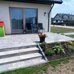 DB.Building Dominik Budzyński Usługi Wypożyczalnia - Nowoczesny taras z kostki brukowej z trzema stopniami, kwiatami w donicy i rabatą kwiatową. W tle dom z dużymi oknami i plac zabaw dla dzieci.