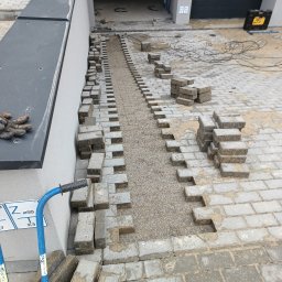 DB.Building Dominik Budzyński Usługi Wypożyczalnia - Układanie szarej kostki brukowej na podsypce z grysu, widoczne narzędzia brukarskie i ułożona część nawierzchni przy budynku z garażem.