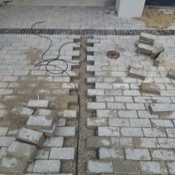 DB.Building Dominik Budzyński Usługi Wypożyczalnia - Układanie szarej kostki brukowej z widocznym korytkiem odwadniającym wypełnionym żwirem. Część kostki jeszcze nieułożona, widoczne przewody i studzienka kanalizacyjna.