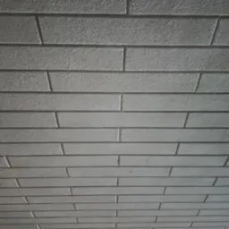 Szara ściana wykończona imitacją cegły, widoczna z dołu pod kątem, z delikatną fakturą i przesuniętymi spoinami.