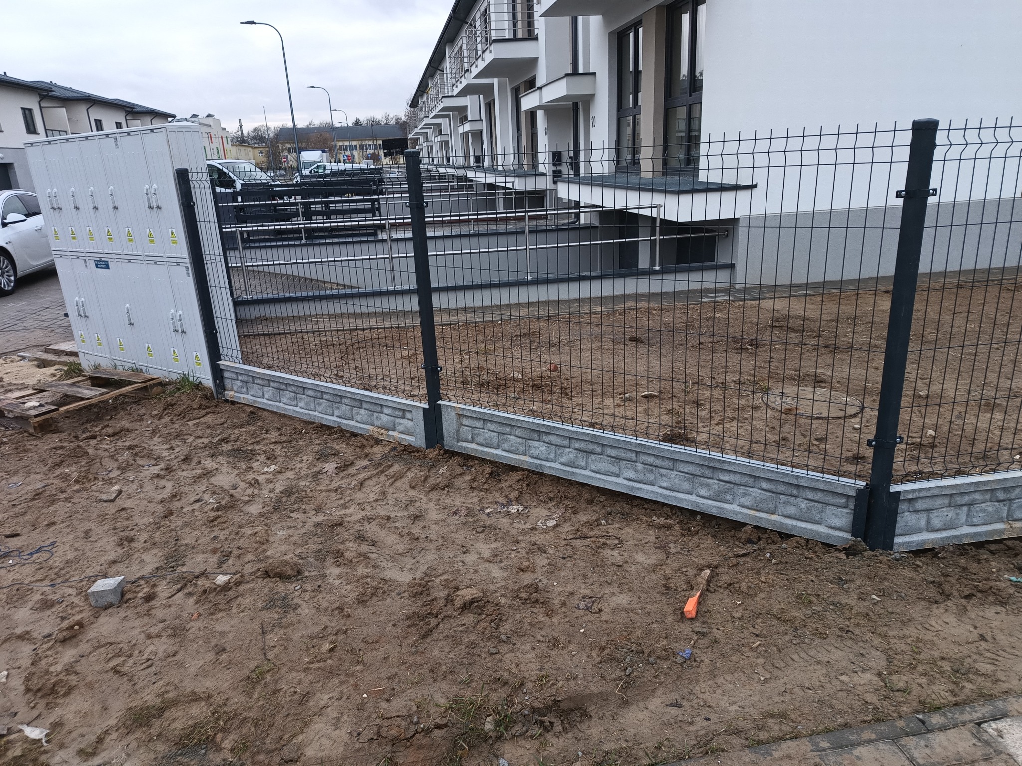 Nowoczesne ogrodzenie panelowe z podmurówką imitującą cegłę, oddzielające teren budowy od nowej zabudowy szeregowej, widoczne skrzynki elektryczne i narzędzie w ziemi.