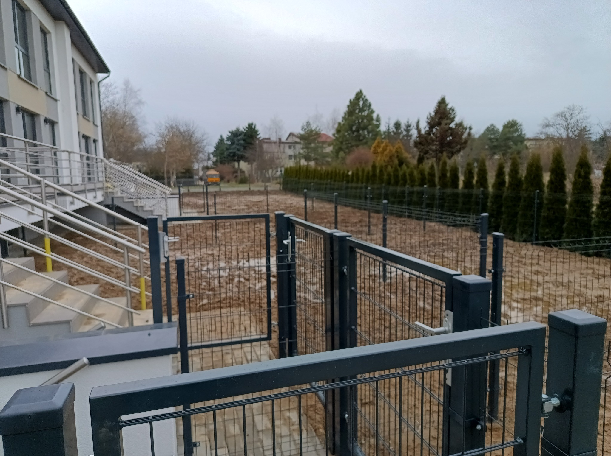 Nowe, grafitowe ogrodzenie panelowe z furtką na posesji, widok z góry na ziemię po deszczu i rząd tuj w tle.