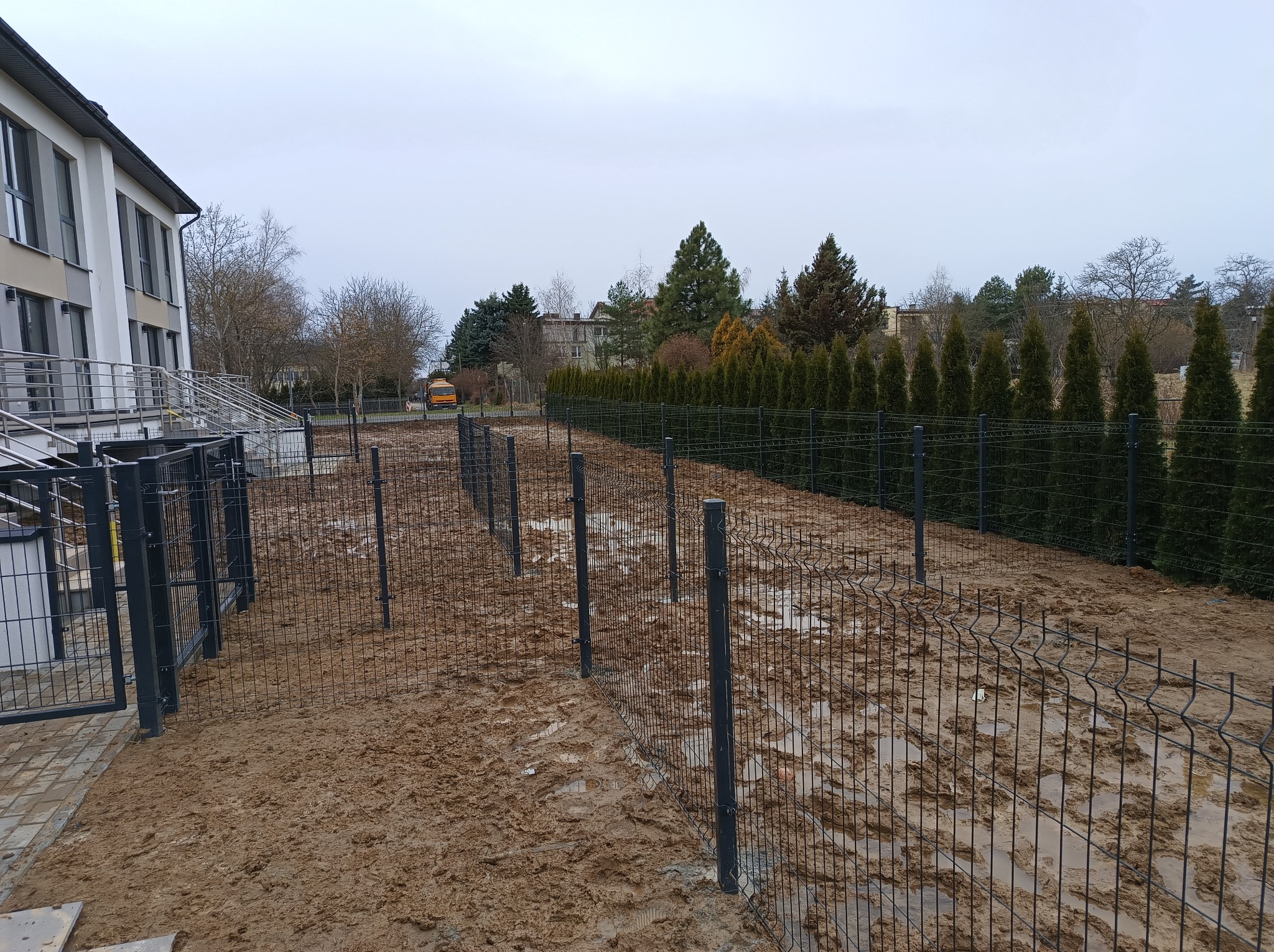 Etap budowy: Czarny metalowy szkielet ogrodzenia panelowego w błotnistym terenie obok budynku z szarymi oknami, w tle rząd wysokich tuj i żółty autobus.