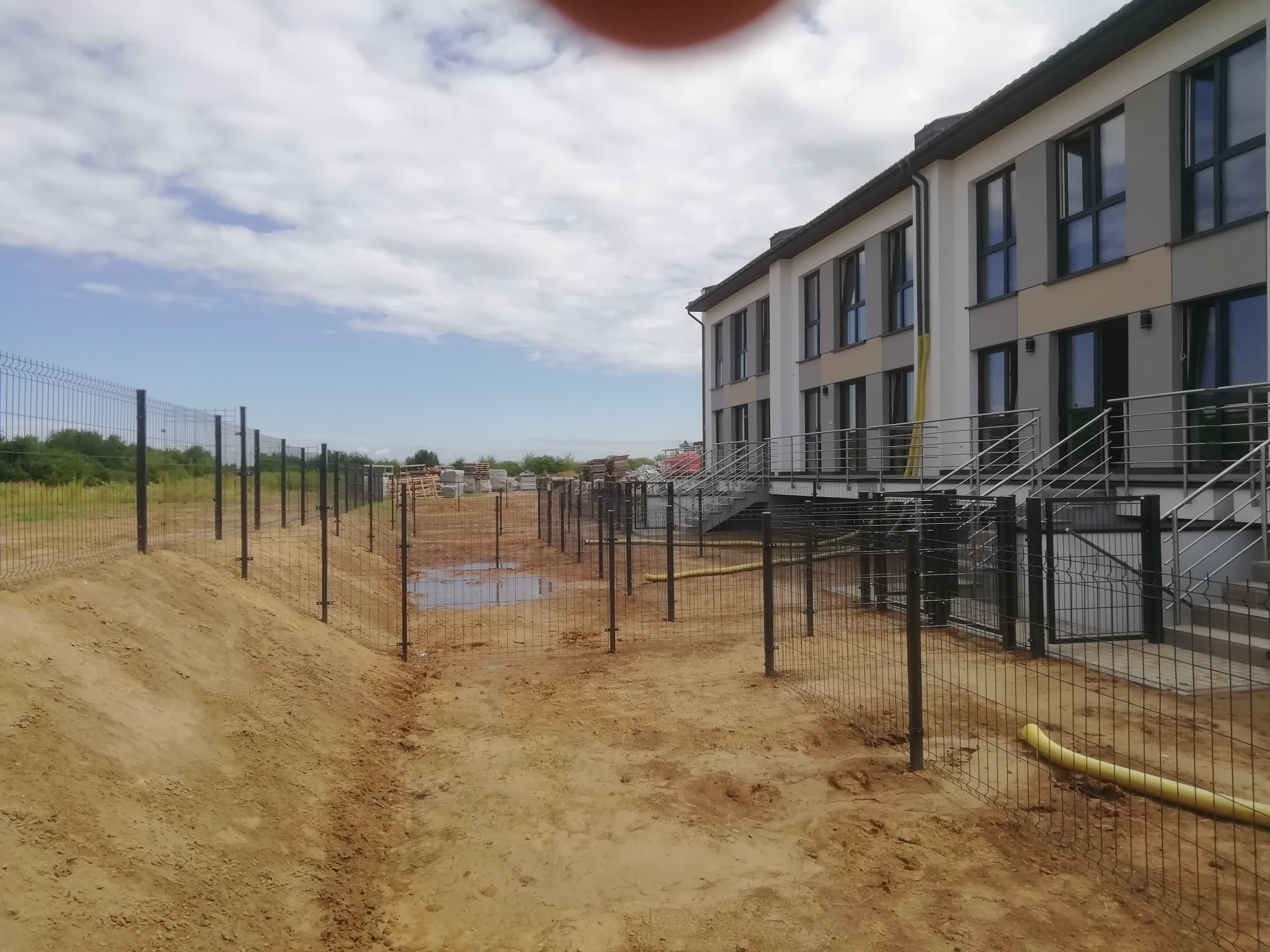 Montaż ogrodzenia panelowego z widocznymi słupkami i częściowo zamontowanymi panelami na nierównym terenie, budynek w tle.