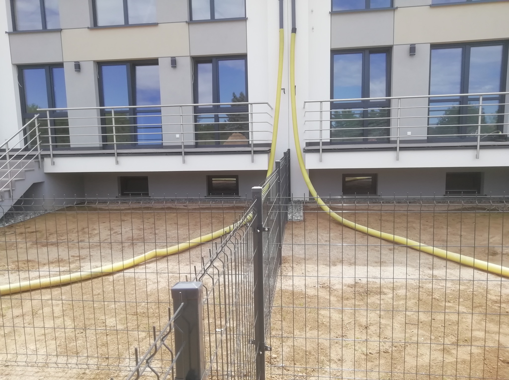 Nowoczesne ogrodzenie panelowe z siatki z widocznymi żółtymi rurami wentylacyjnymi na tle budynku mieszkalnego w trakcie budowy.