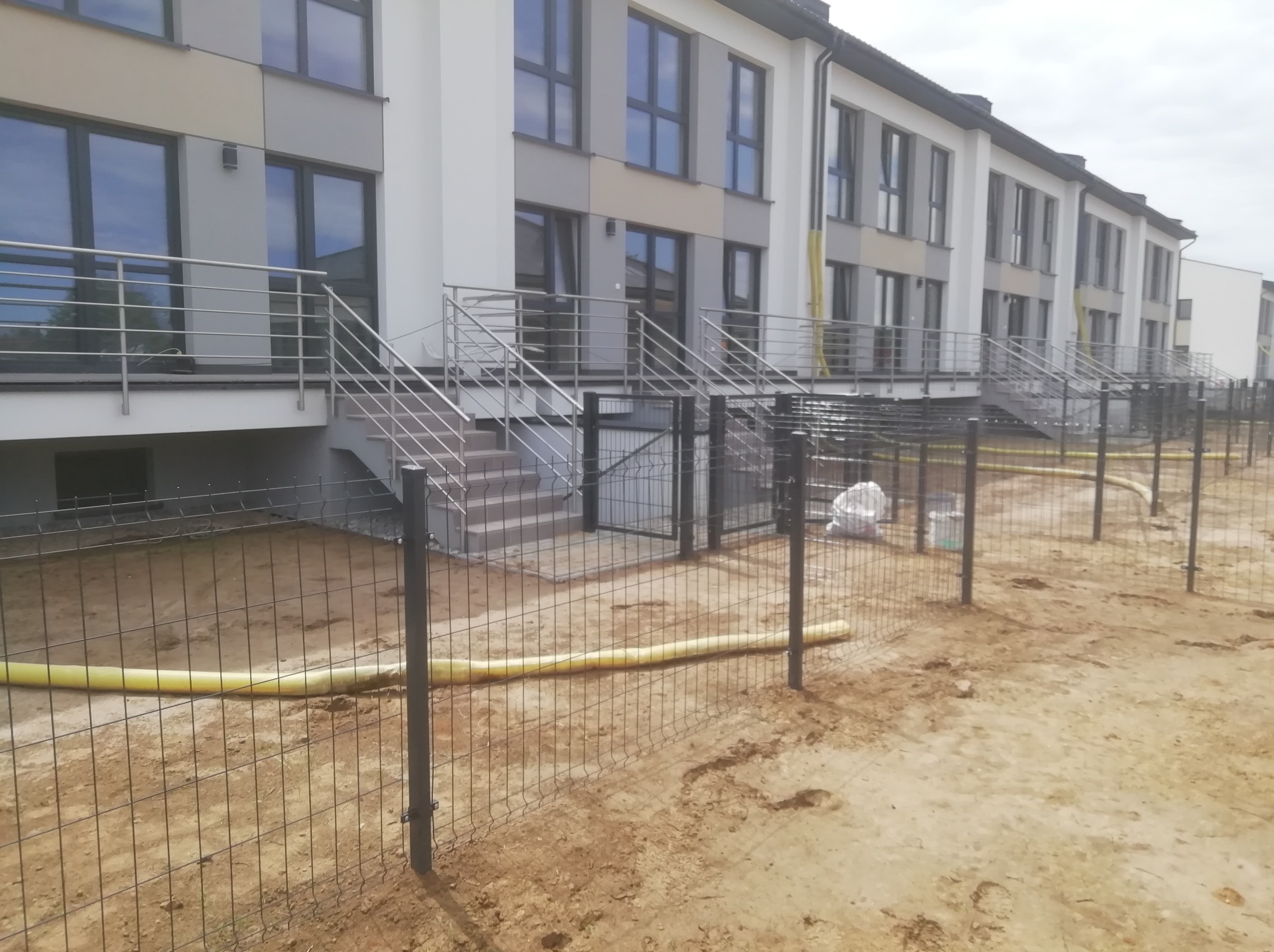 Nowoczesne segmentowe domy w zabudowie szeregowej z metalowym ogrodzeniem z siatki, widoczne żółte rury na ziemi i materiały budowlane w workach.