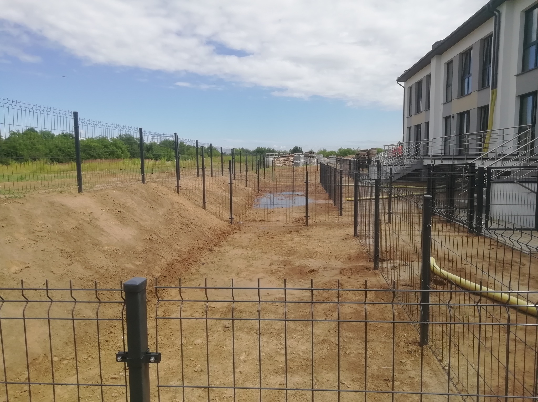 Etap budowy ogrodzenia panelowego z metalową siatką na terenie budowy, widoczne słupki i częściowo zamontowane panele wzdłuż wykopu, w tle budynek mieszkalny i składowane materiały budowlane.