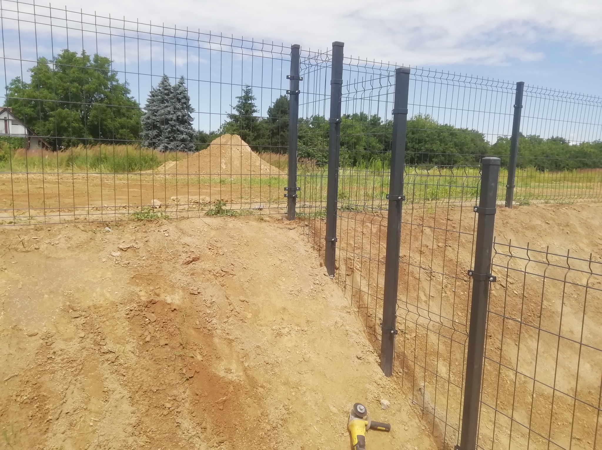 Wykop pod czarne słupki i metalowe panele tworzące częściowo ukończone ogrodzenie na nierównym terenie, z widocznym narzędziem do cięcia w pobliżu.