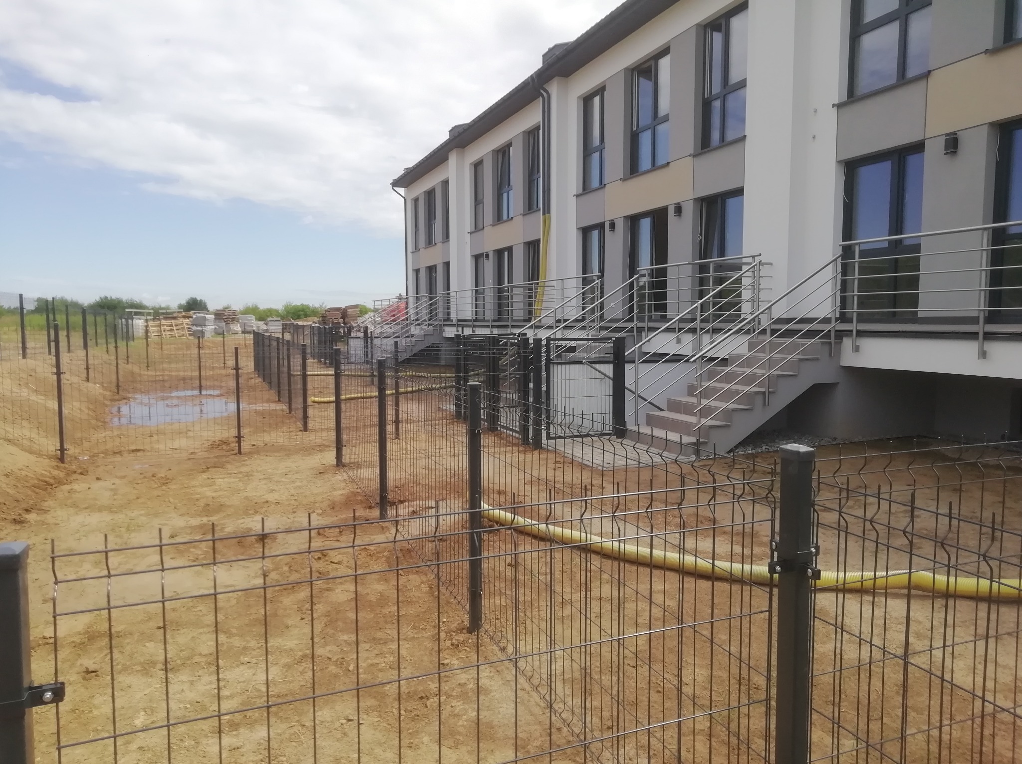 Wznoszone ogrodzenie panelowe z metalowej siatki na nowym osiedlu, widoczne słupki i częściowo zamontowane panele, w tle budynki z metalowymi schodami i żółta rura na ziemi.