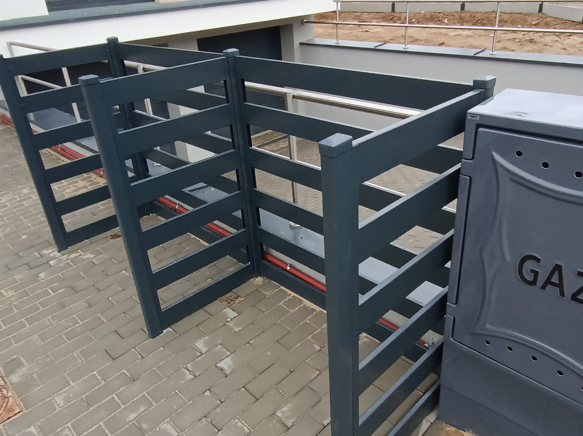 Szare, ażurowe ogrodzenie śmietnika z drewnopodobnych paneli na kostce brukowej, z widoczną skrzynką gazową po prawej stronie.