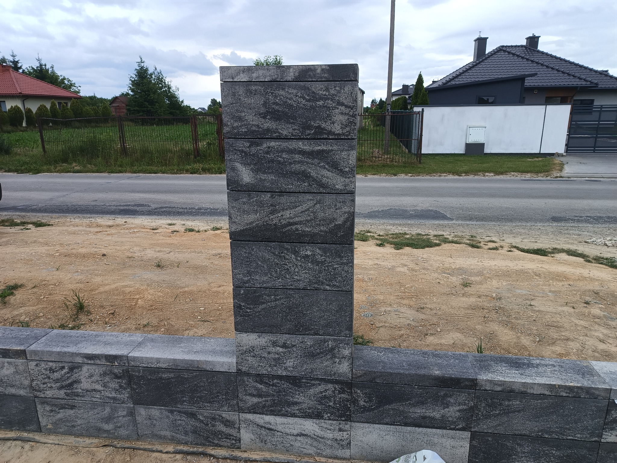 Fragment ogrodzenia betonowego z szarej kostki, słupek i podmurówka, na tle drogi i zabudowań. Widoczne nierówności terenu.