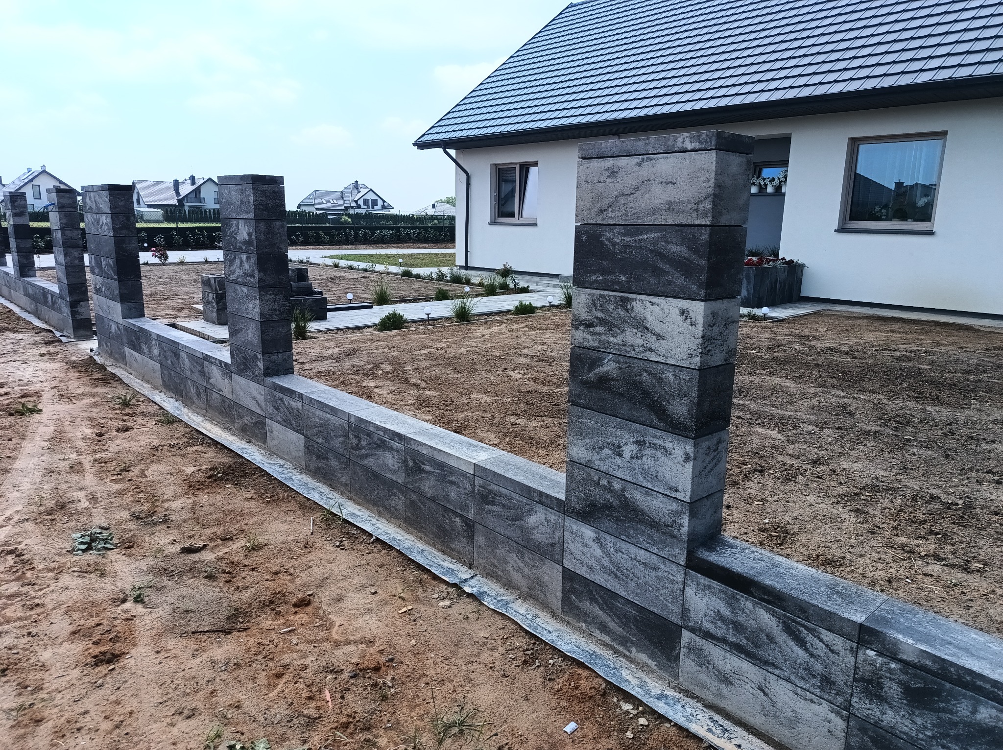 Wznoszenie ogrodzenia z szarych, betonowych bloczków. Widoczne słupki i fragment murku, w tle dom jednorodzinny i trawnik. Prace w toku, teren wokół ogrodzenia niezagospodarowany.