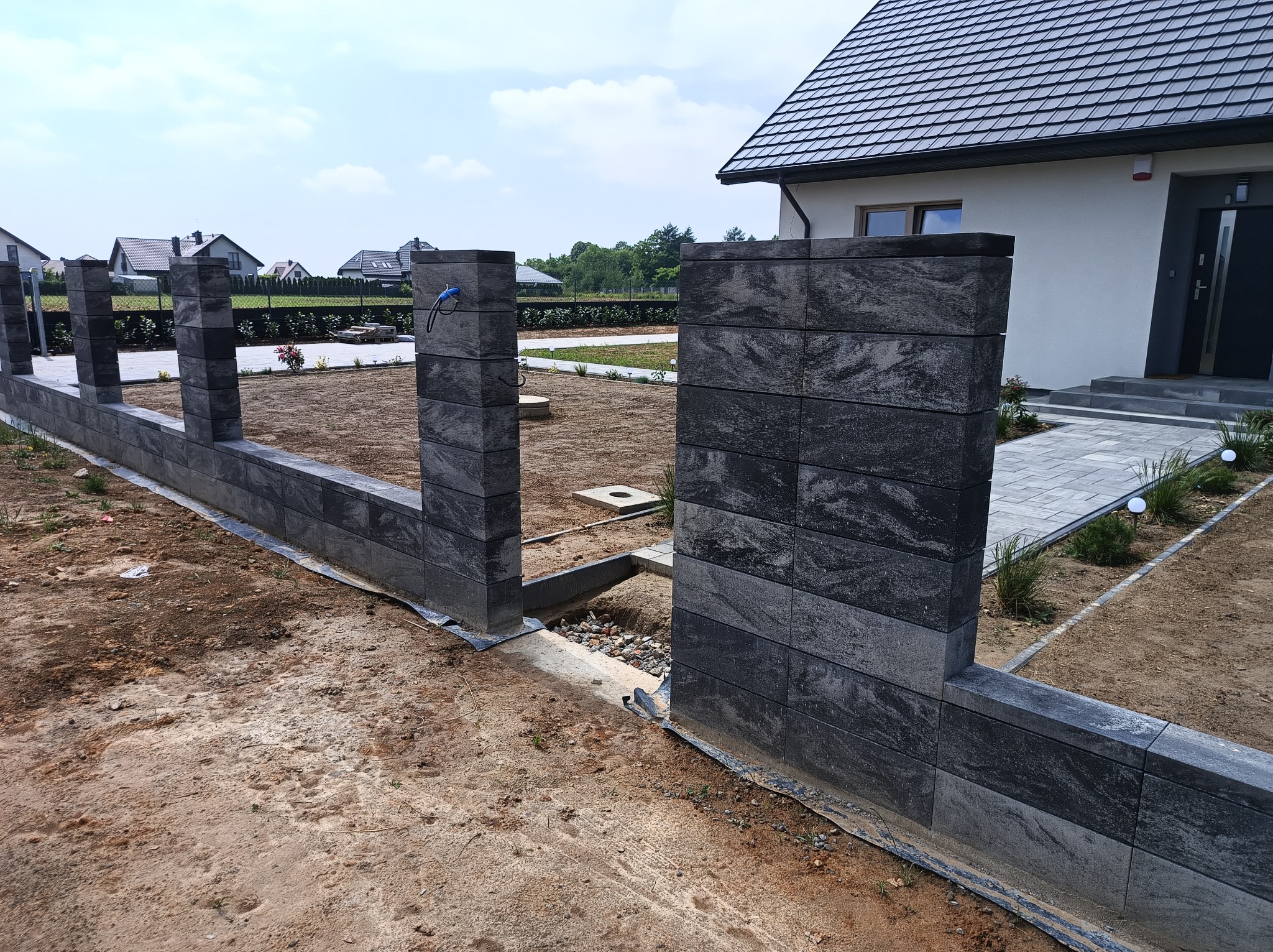 Nowoczesne ogrodzenie z szarej kostki betonowej, widok z bliska na fragment z przerwą, w tle dom z szarą stolarką i kostką brukową.