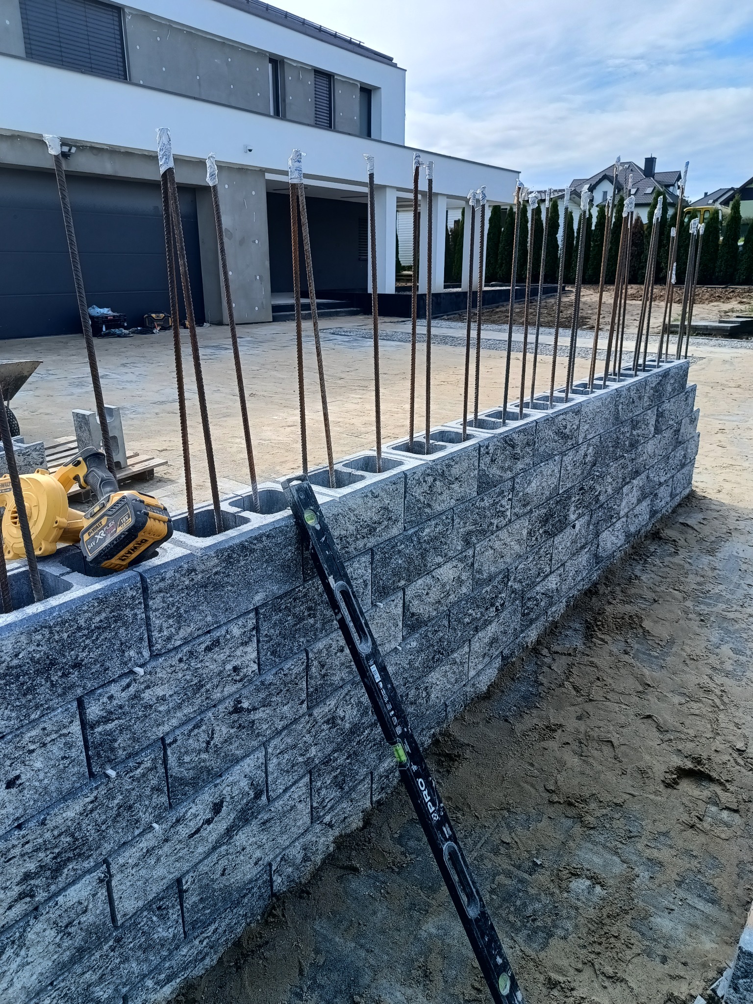 Wznoszenie muru z szarych betonowych bloczków z wystającymi prętami zbrojeniowymi, poziomica i narzędzia obok, nowoczesny dom w tle, etap budowy ogrodzenia.