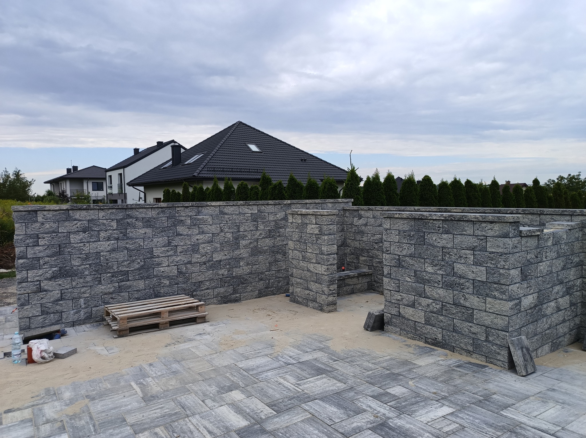Szara, częściowo ukończona konstrukcja z bloczków betonowych na posesji z kostką brukową, widok na domy i pochmurne niebo w tle.