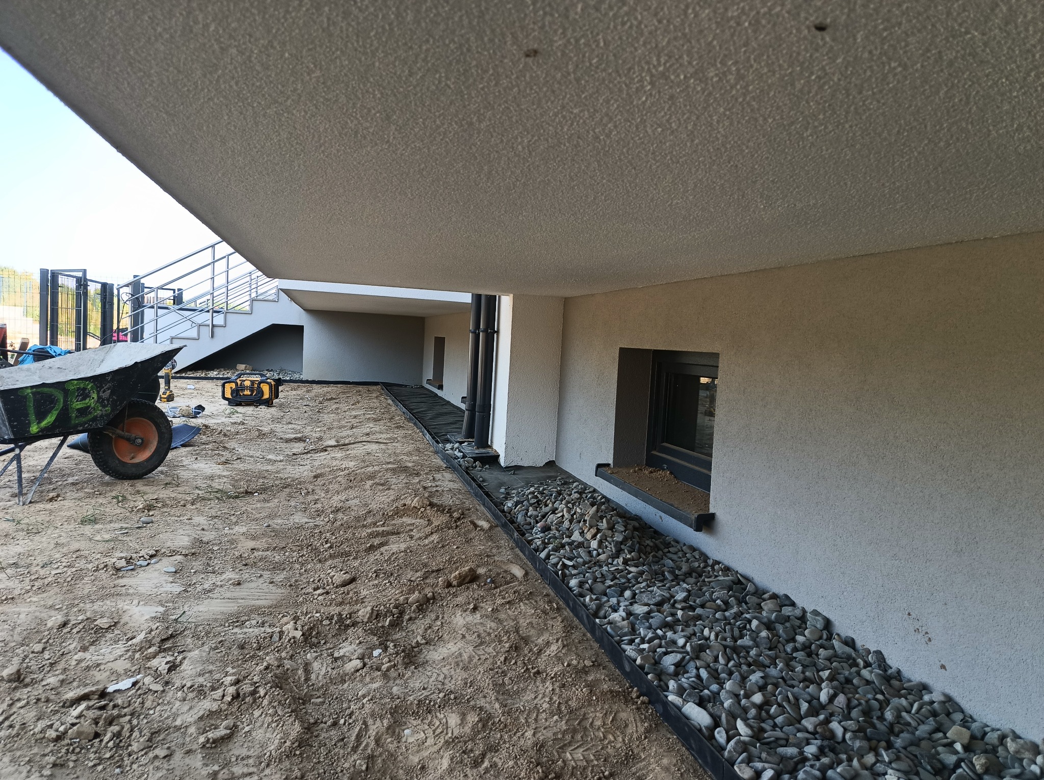 Wykończona elewacja budynku z drenażem z kamieni, widok z dołu. Taczka i narzędzia budowlane w tle. Nowoczesne wykończenie.