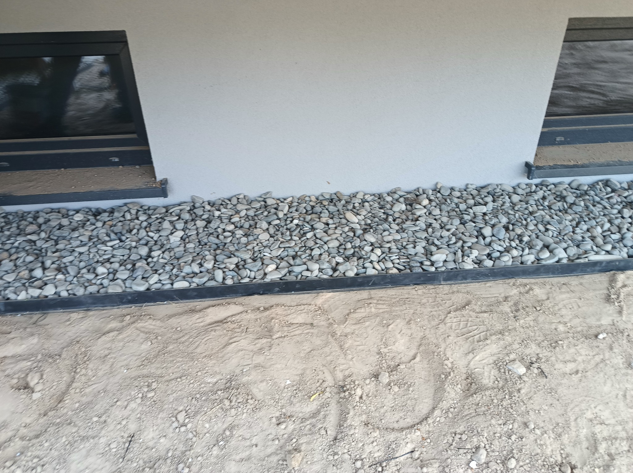 Wykończenie elewacji budynku z szarymi kamieniami przy ścianie i oknach piwnicznych. Widoczny fragment ściany i ziemia. Estetyczne detale.