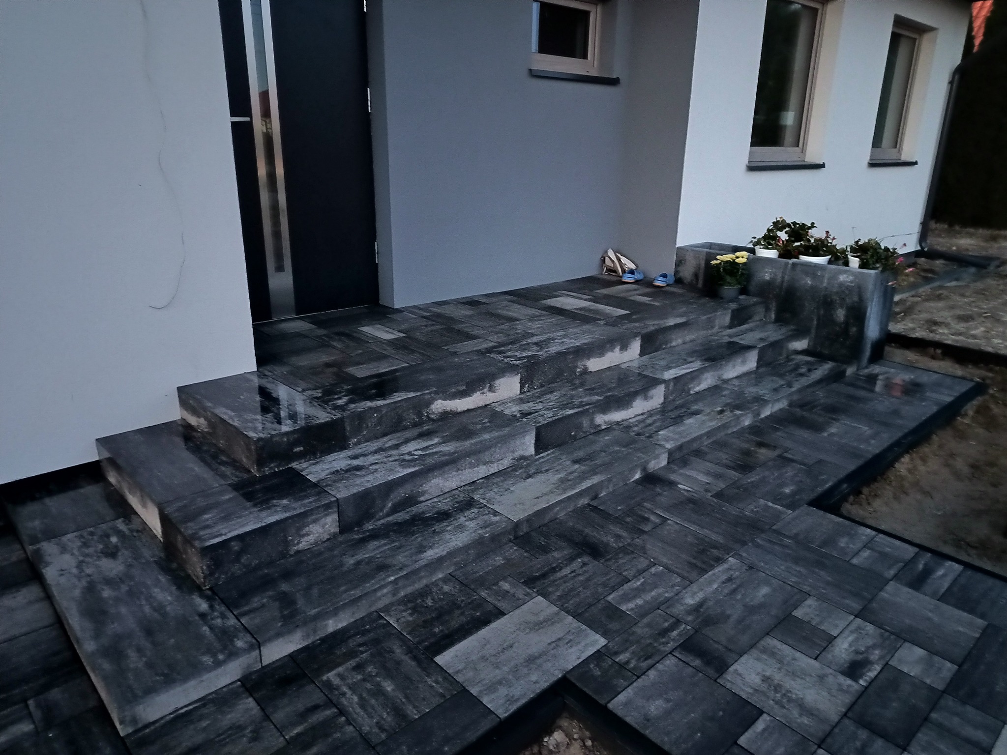 Schody wejściowe wyłożone ciemną kostką brukową o nieregularnym wzorze, prowadzące do nowoczesnego domu z szarą elewacją i czarnymi drzwiami.