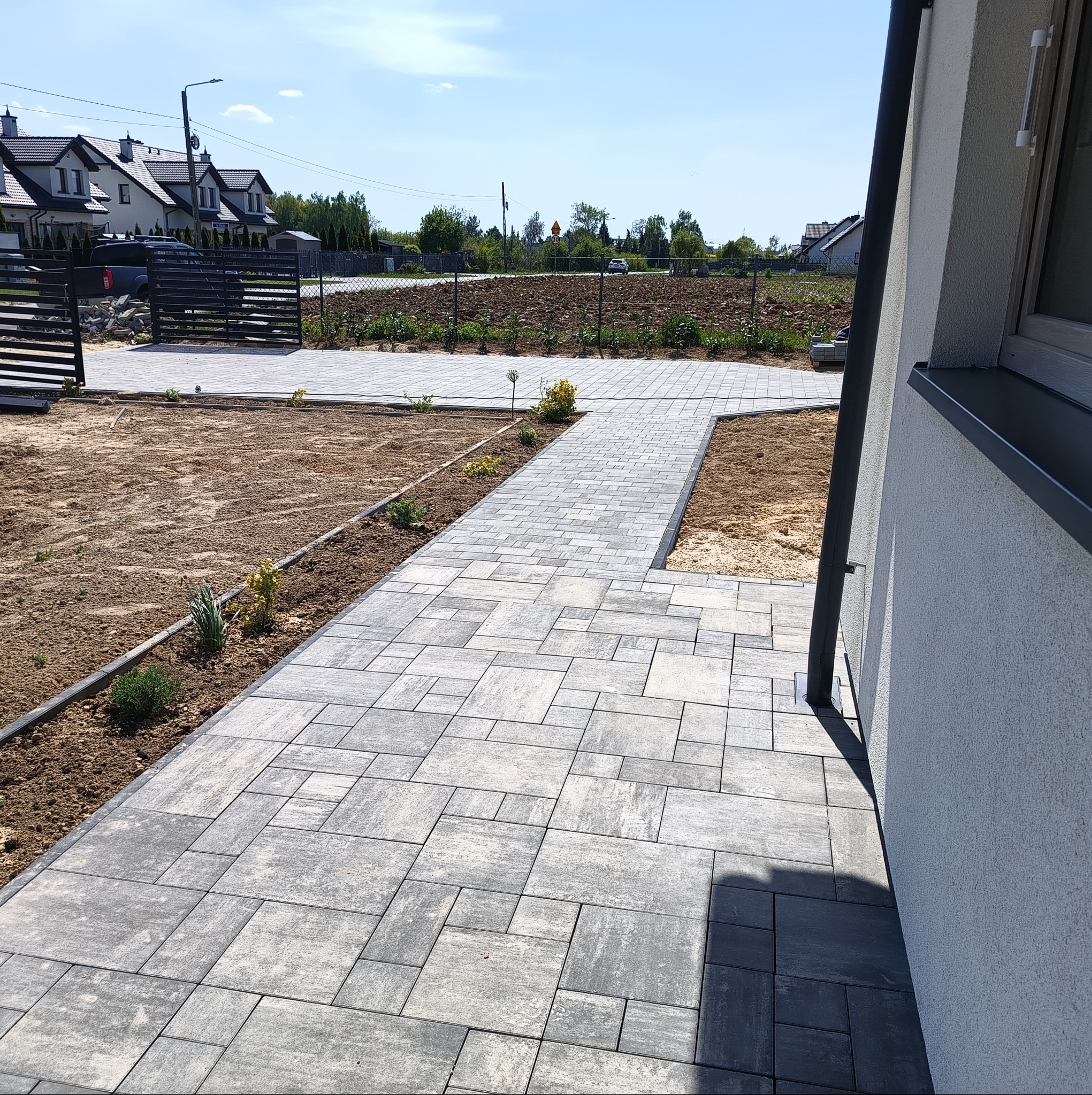 Ułożona kostka brukowa tworząca ścieżkę w ogrodzie, z widocznymi rabatami i fragmentem domu. Jasne, dzienne światło podkreśla teksturę kostki.