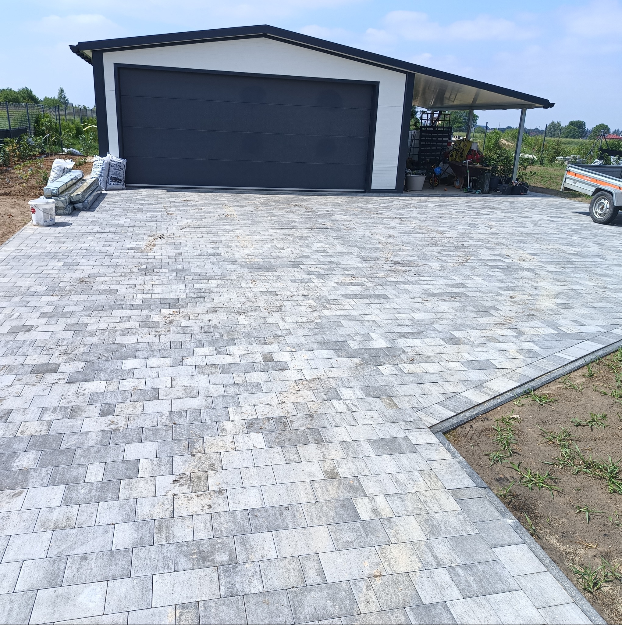 Ułożona szara kostka brukowa na podjeździe, z widocznym garażem w tle. Kostka ułożona w regularny wzór, obrzeża wykończone. Częściowo widoczne worki z materiałami.