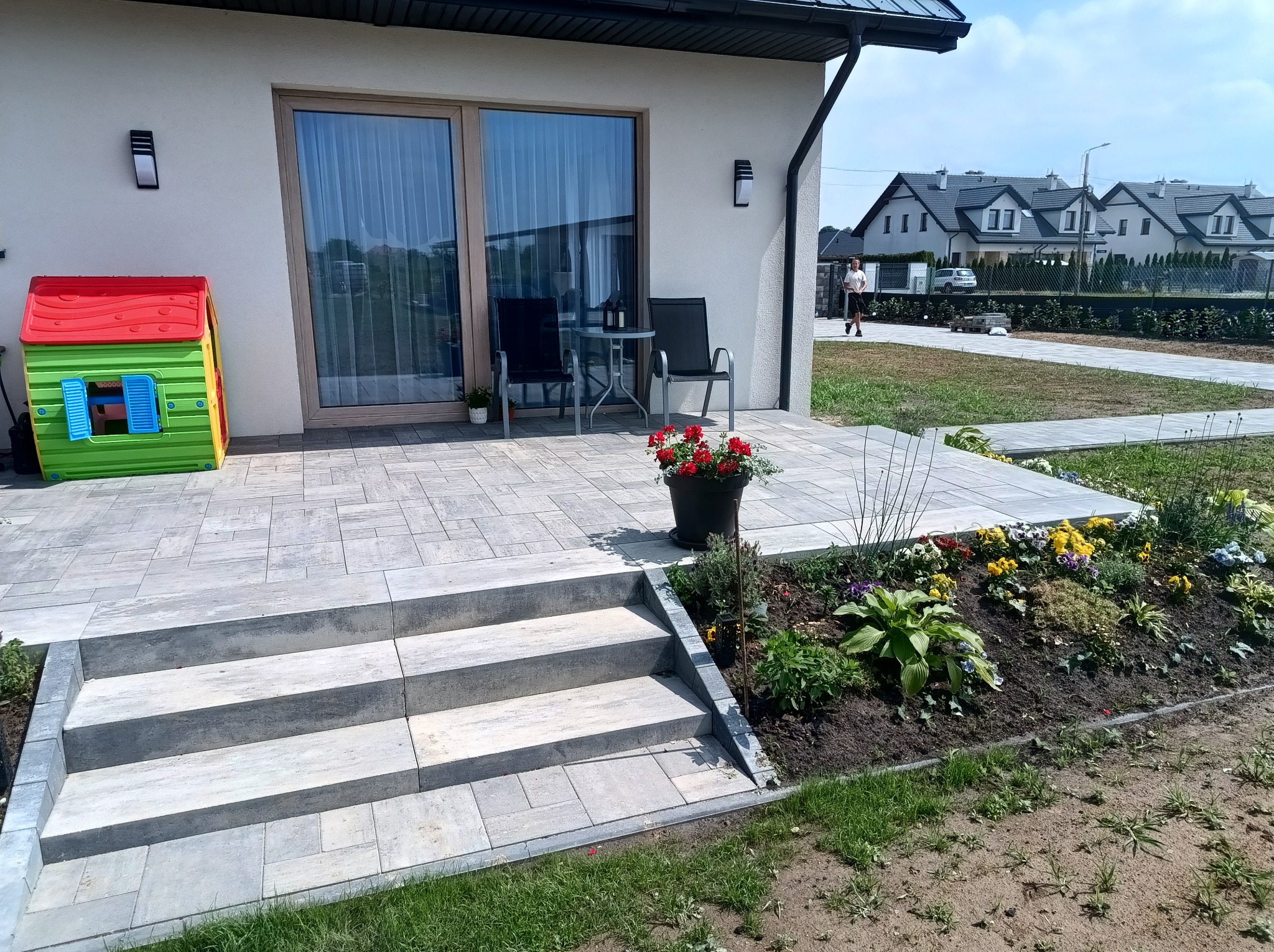 Nowoczesny taras z kostki brukowej z trzema stopniami, kwiatami w donicy i rabatą kwiatową. W tle dom z dużymi oknami i plac zabaw dla dzieci.