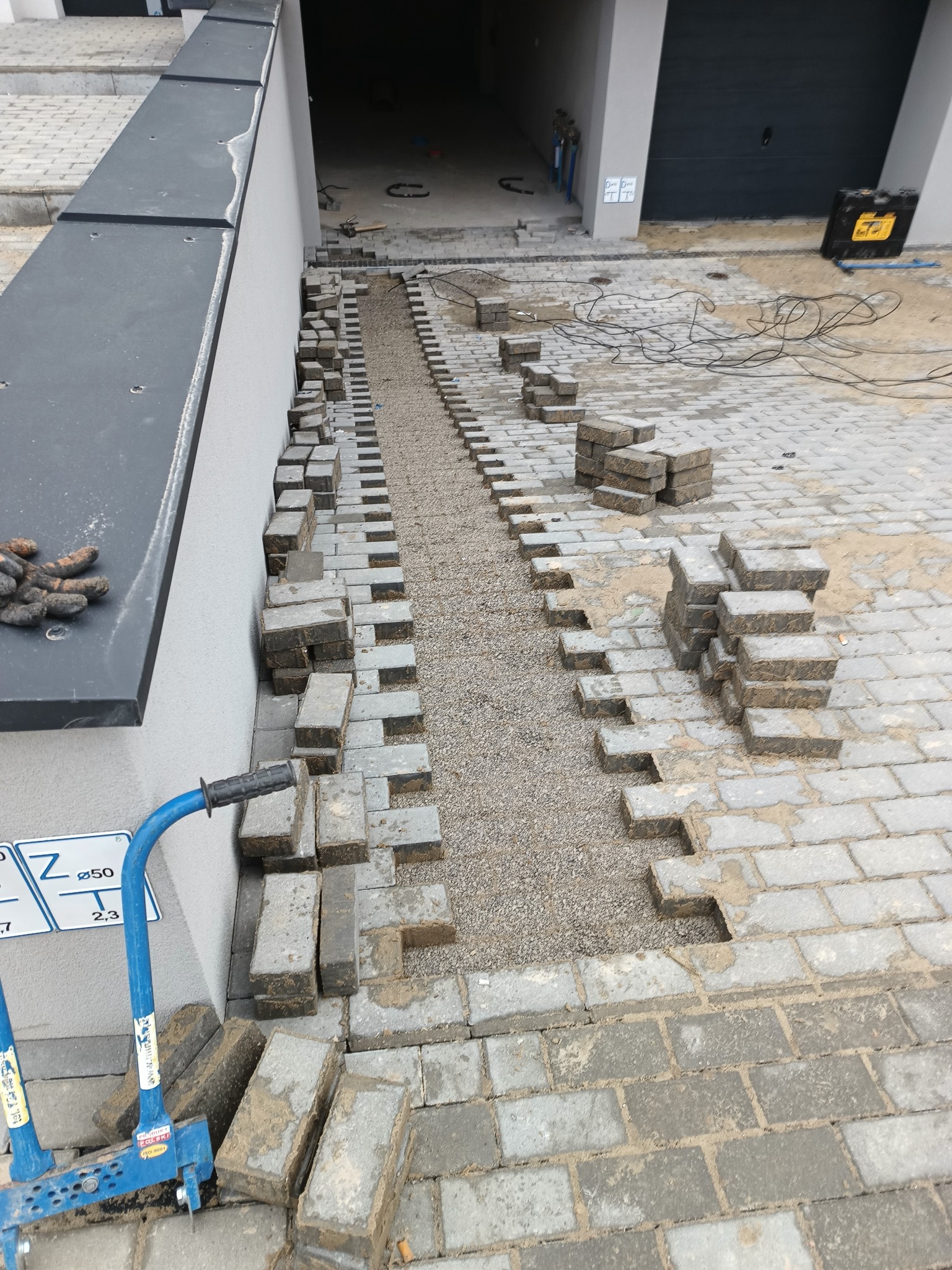 Układanie szarej kostki brukowej na podsypce z grysu, widoczne narzędzia brukarskie i ułożona część nawierzchni przy budynku z garażem.