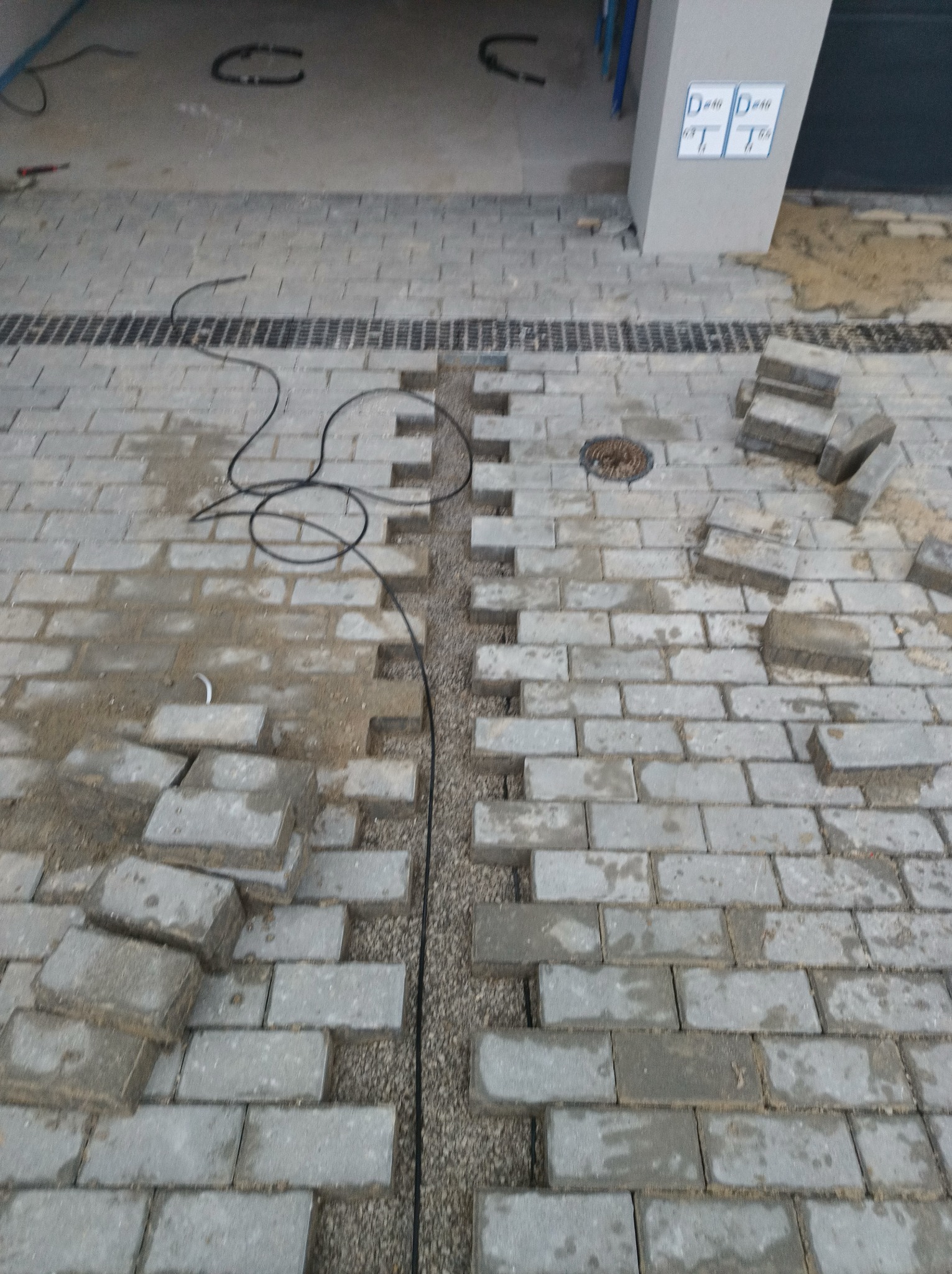 Układanie szarej kostki brukowej z widocznym korytkiem odwadniającym wypełnionym żwirem. Część kostki jeszcze nieułożona, widoczne przewody i studzienka kanalizacyjna.