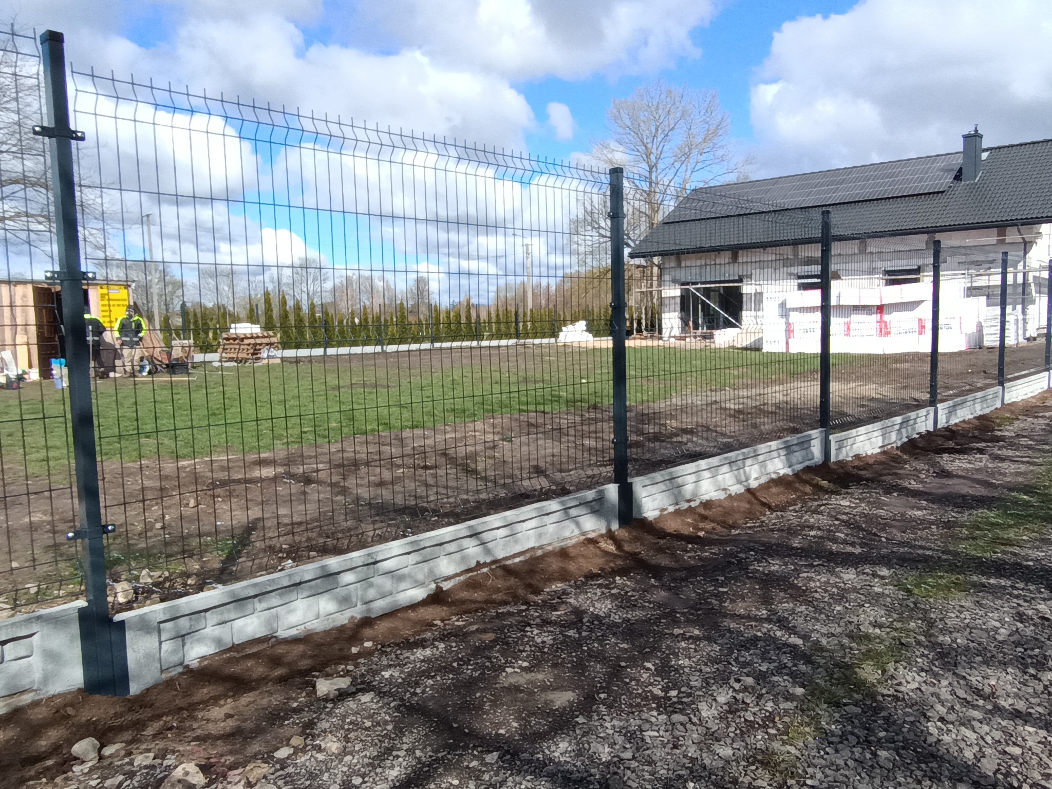 Montaż ogrodzenia panelowego na fundamencie z prefabrykatów betonowych. W tle budynek w trakcie budowy i pracownicy. Widok z zewnątrz posesji.