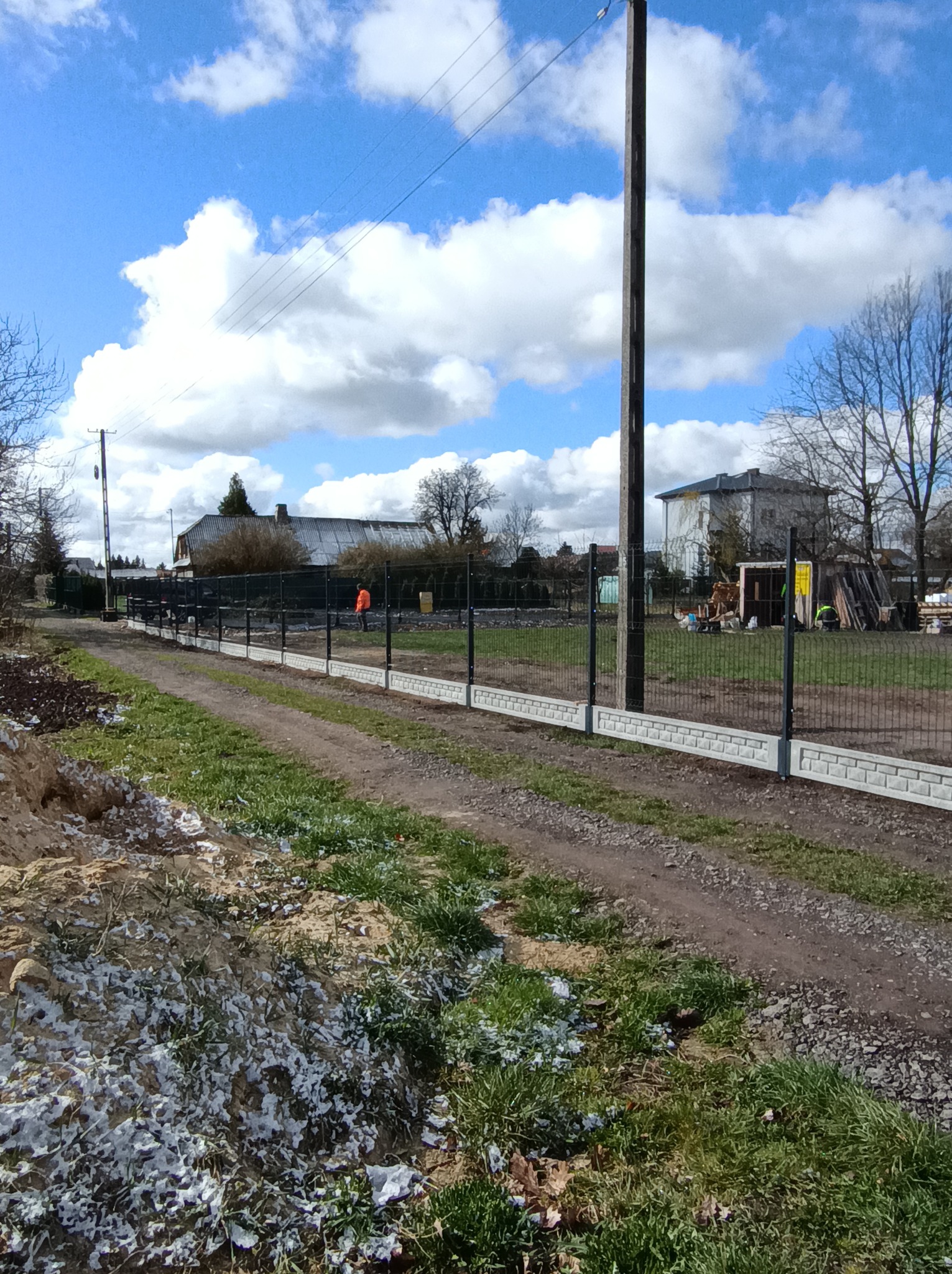 Nowoczesne ogrodzenie panelowe z podmurówką imitującą cegłę, otoczone zielenią i ziemią. W tle budynki mieszkalne pod błękitnym niebem z chmurami.