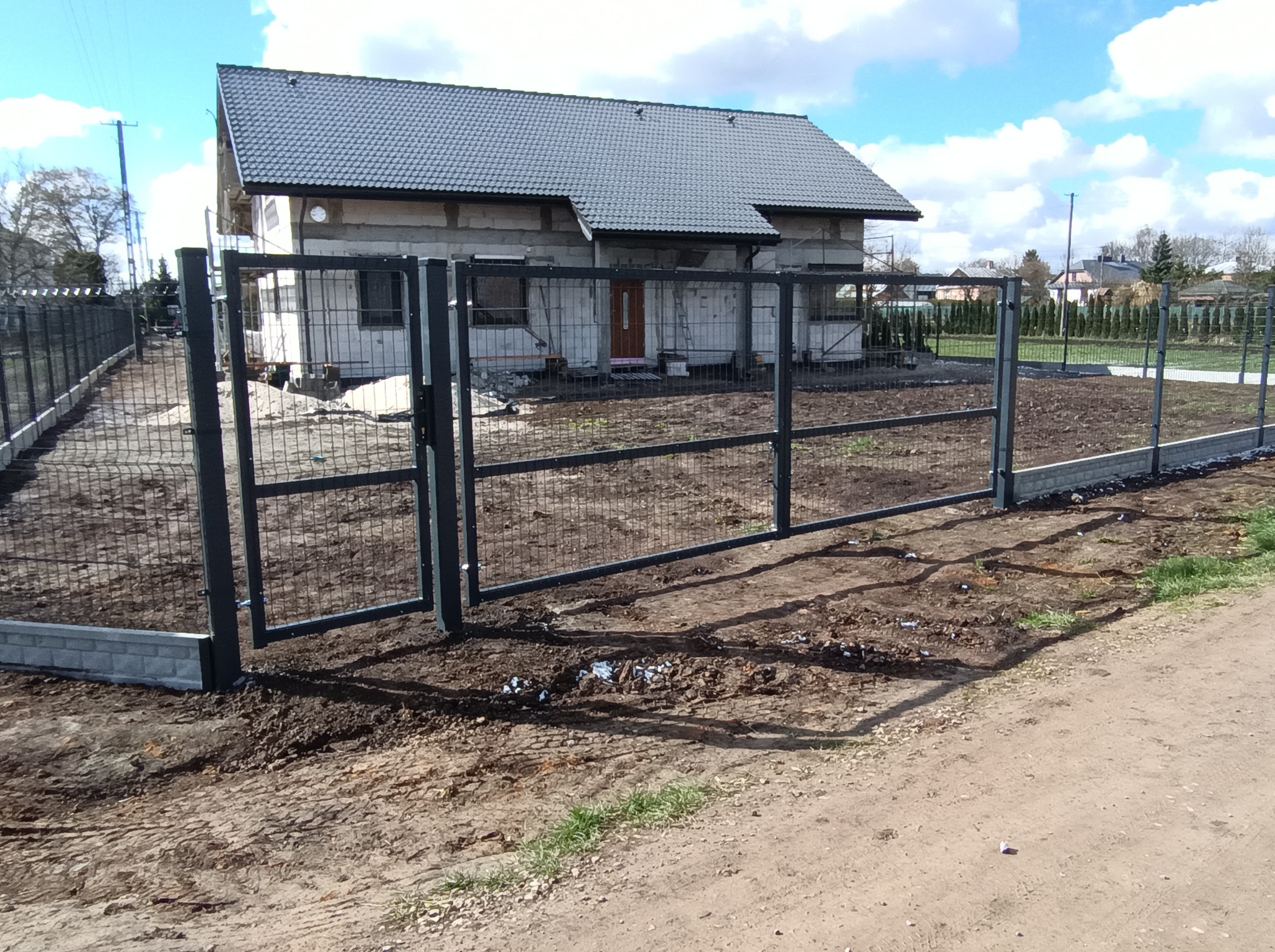 Nowoczesne, grafitowe ogrodzenie panelowe z bramą wjazdową na tle domu w trakcie budowy. Widoczne słupki i podmurówka z szarej kostki. Słoneczny dzień.