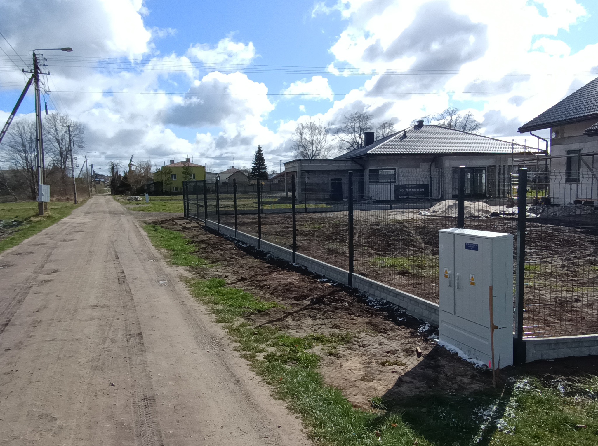 Nowoczesne ogrodzenie panelowe z podmurówką na nierównym terenie, skrzynka elektryczna obok, w tle dom w budowie i polna droga. Słoneczny dzień.