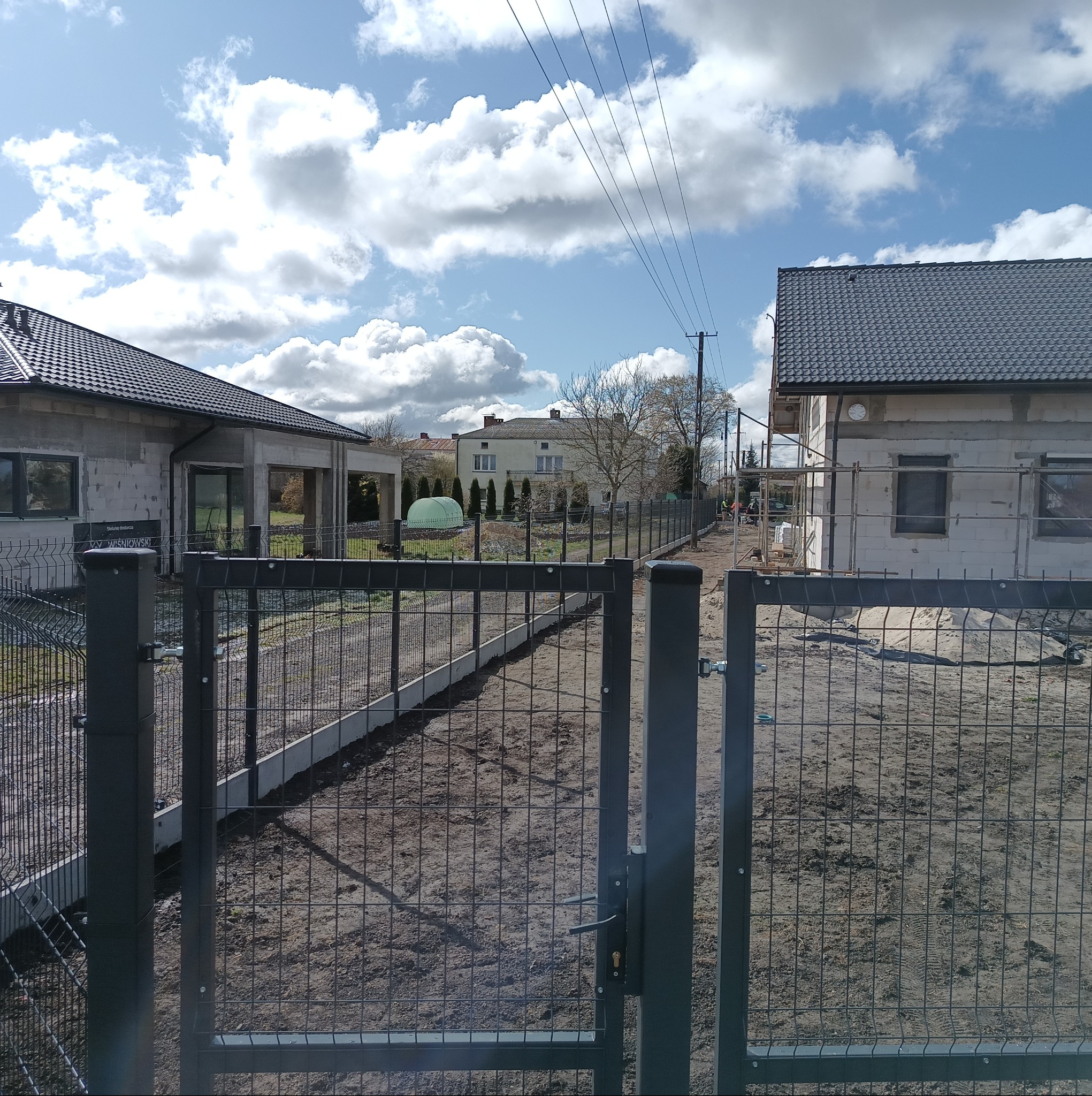 Nowoczesne ogrodzenie panelowe w kolorze grafitowym, z widoczną furtką i betonową podmurówką, otaczające posesję z domami w trakcie budowy, na tle błękitnego nieba.