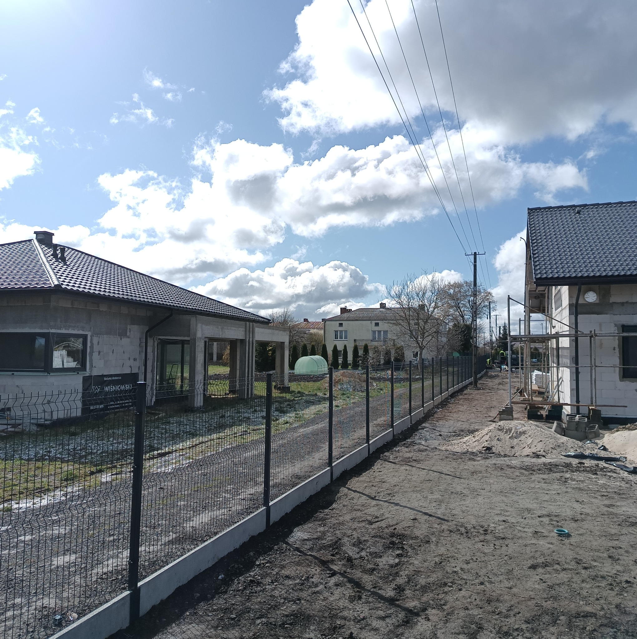 Nowoczesne ogrodzenie panelowe z podmurówką betonową, widoczne na tle budynków w trakcie budowy, w słoneczny dzień. Elementy montażowe i teren budowy.