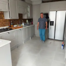 Jasnoszara kuchnia po remoncie z ceglaną ścianą, jasną podłogą i mężczyzną w paski stojącym na środku, widoczne środki czystości na podłodze.