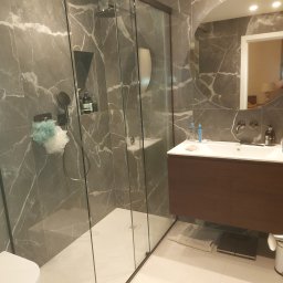 Piotr Wolski - Nowoczesna łazienka z kabiną prysznicową typu walk-in, marmurowymi płytkami w odcieniach szarości, wiszącą szafką z umywalką i okrągłym lustrem z podświetleniem, uzupełniona o białą toaletę...