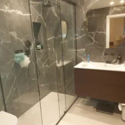 Nowoczesna łazienka z kabiną prysznicową typu walk-in, marmurowymi płytkami w odcieniach szarości, wiszącą szafką z umywalką i okrągłym lustrem z podświetleniem, uzupełniona o białą toaletę...