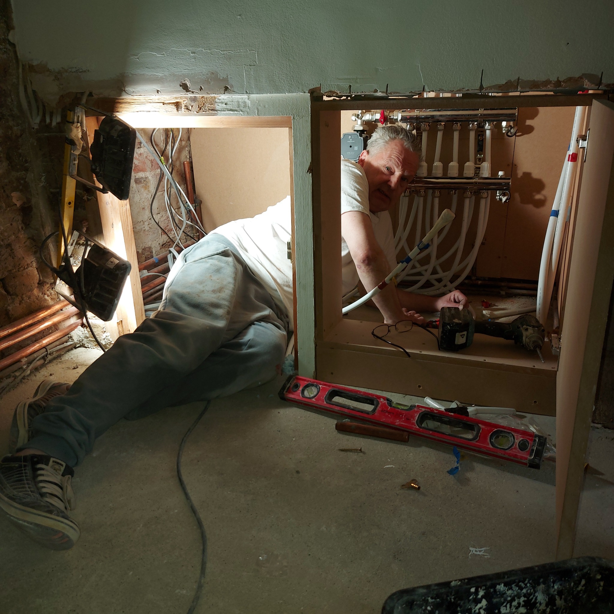 Instalator w trakcie montażu rozdzielacza ogrzewania podłogowego w szafce, widoczne rury, poziomica, wiertarka i narzędzia.