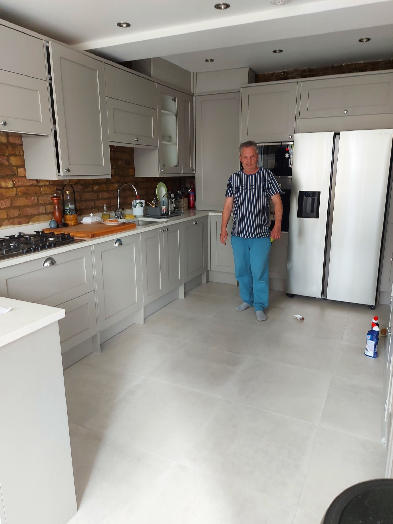 Jasnoszara kuchnia po remoncie z ceglaną ścianą, jasną podłogą i mężczyzną w paski stojącym na środku, widoczne środki czystości na podłodze.