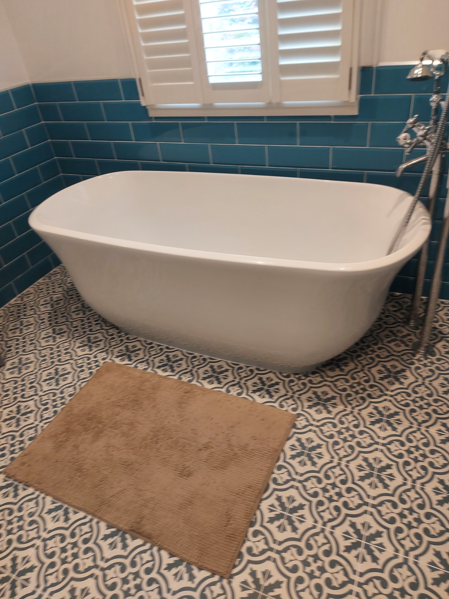 Wolnostojąca wanna z chromowaną armaturą na tle turkusowej ściany z płytek i wzorzystej podłogi, z brązowym dywanikiem łazienkowym.