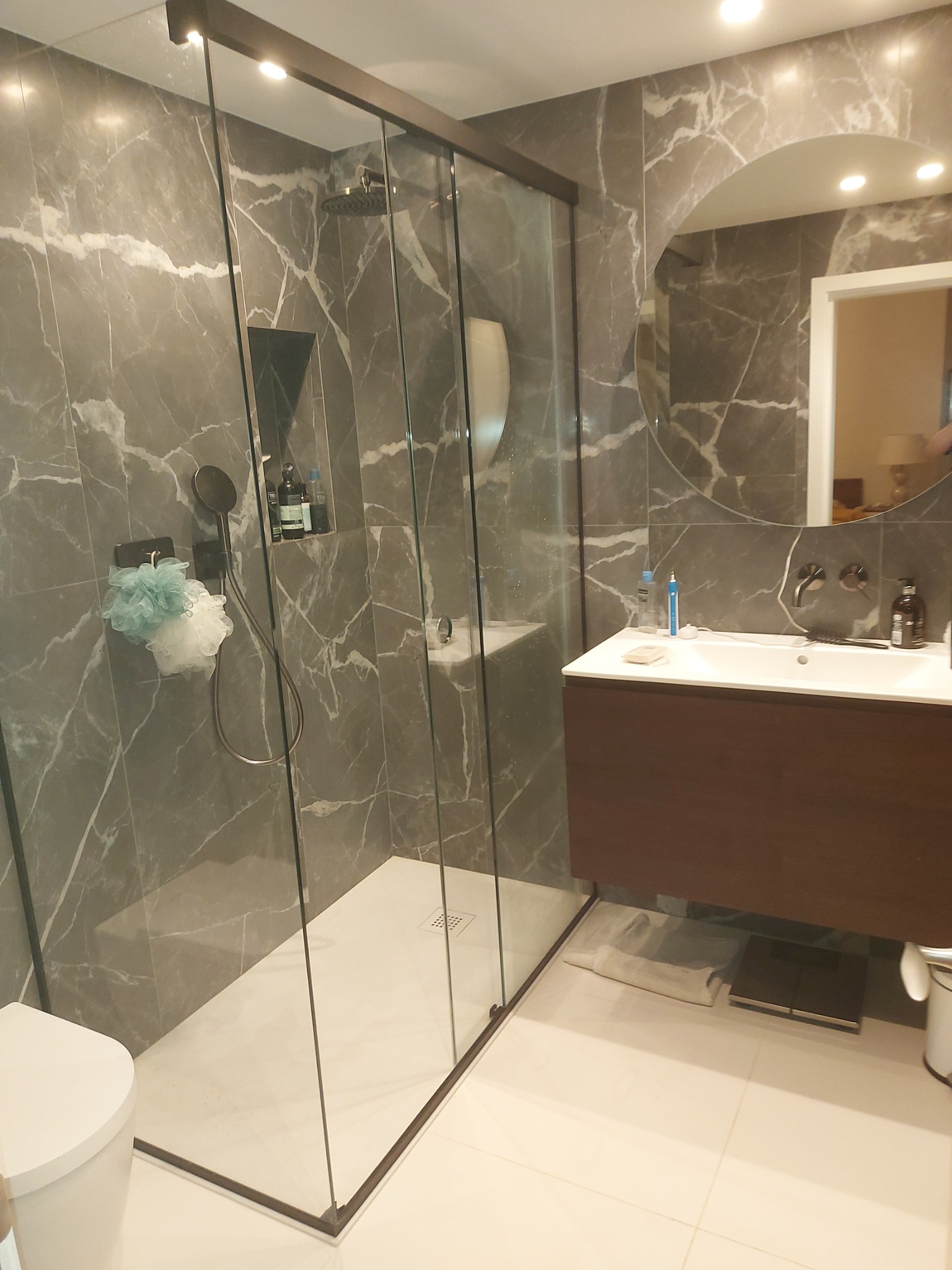 Nowoczesna łazienka z kabiną prysznicową typu walk-in, marmurowymi płytkami w odcieniach szarości, wiszącą szafką z umywalką i okrągłym lustrem z podświetleniem, uzupełniona o białą toaletę...