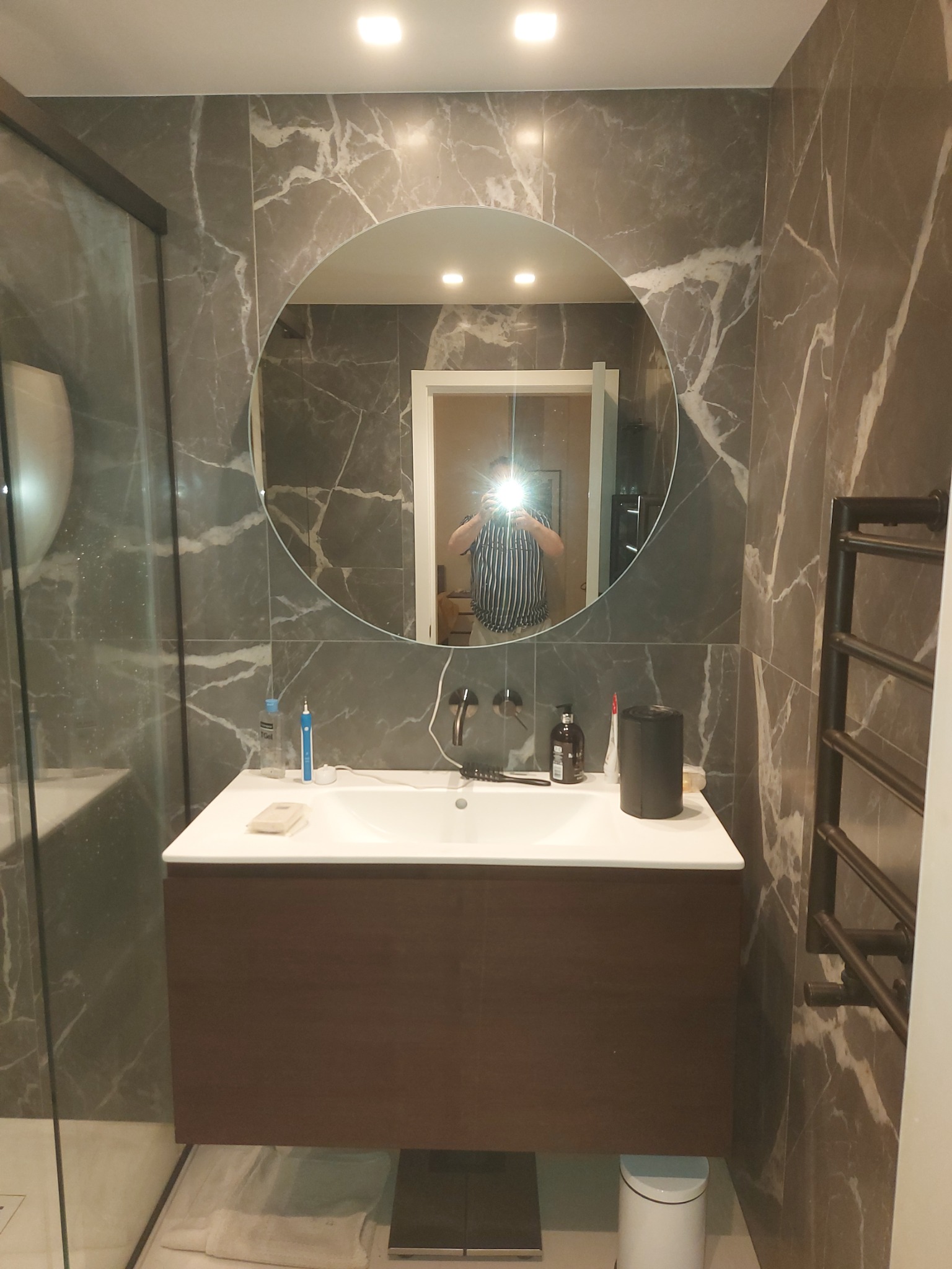 Wykończona łazienka z marmurowymi płytkami, okrągłym lustrem i wiszącą szafką z umywalką, widoczny grzejnik drabinkowy i kabina prysznicowa.