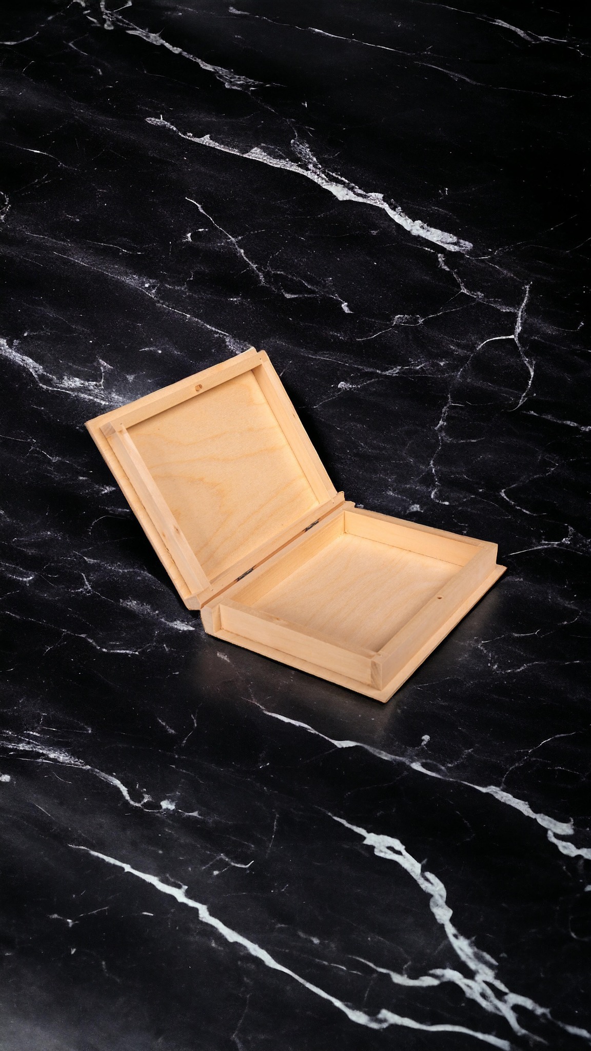 Otwarta, pusta, kwadratowa, drewniana szkatułka z jasnego drewna na czarnym tle imitującym marmur.