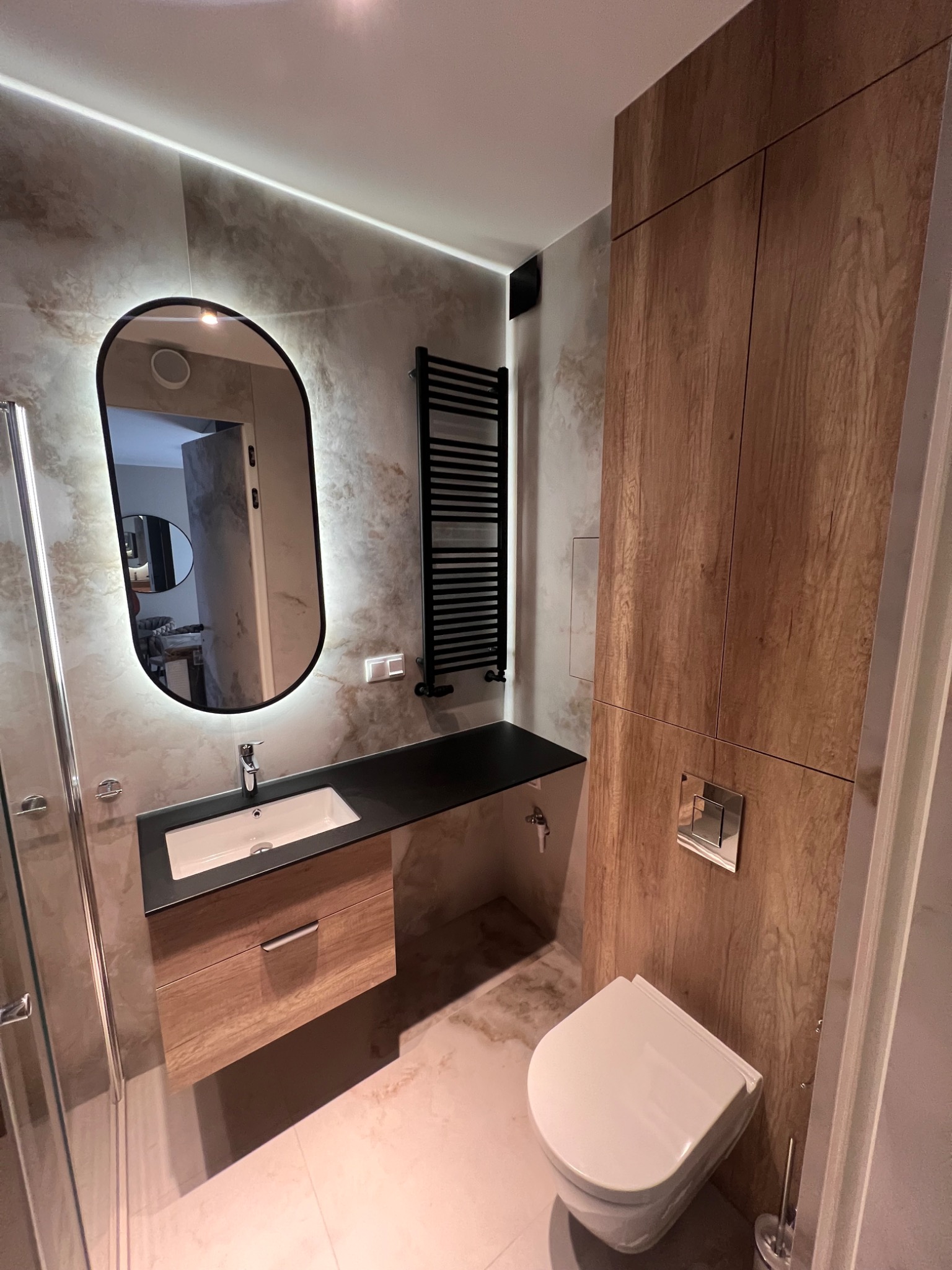 Nowoczesna łazienka po remoncie: owalne lustro z podświetleniem LED, umywalka z szafką, drewnopodobna zabudowa, grzejnik drabinkowy i toaleta w minimalistycznym stylu.