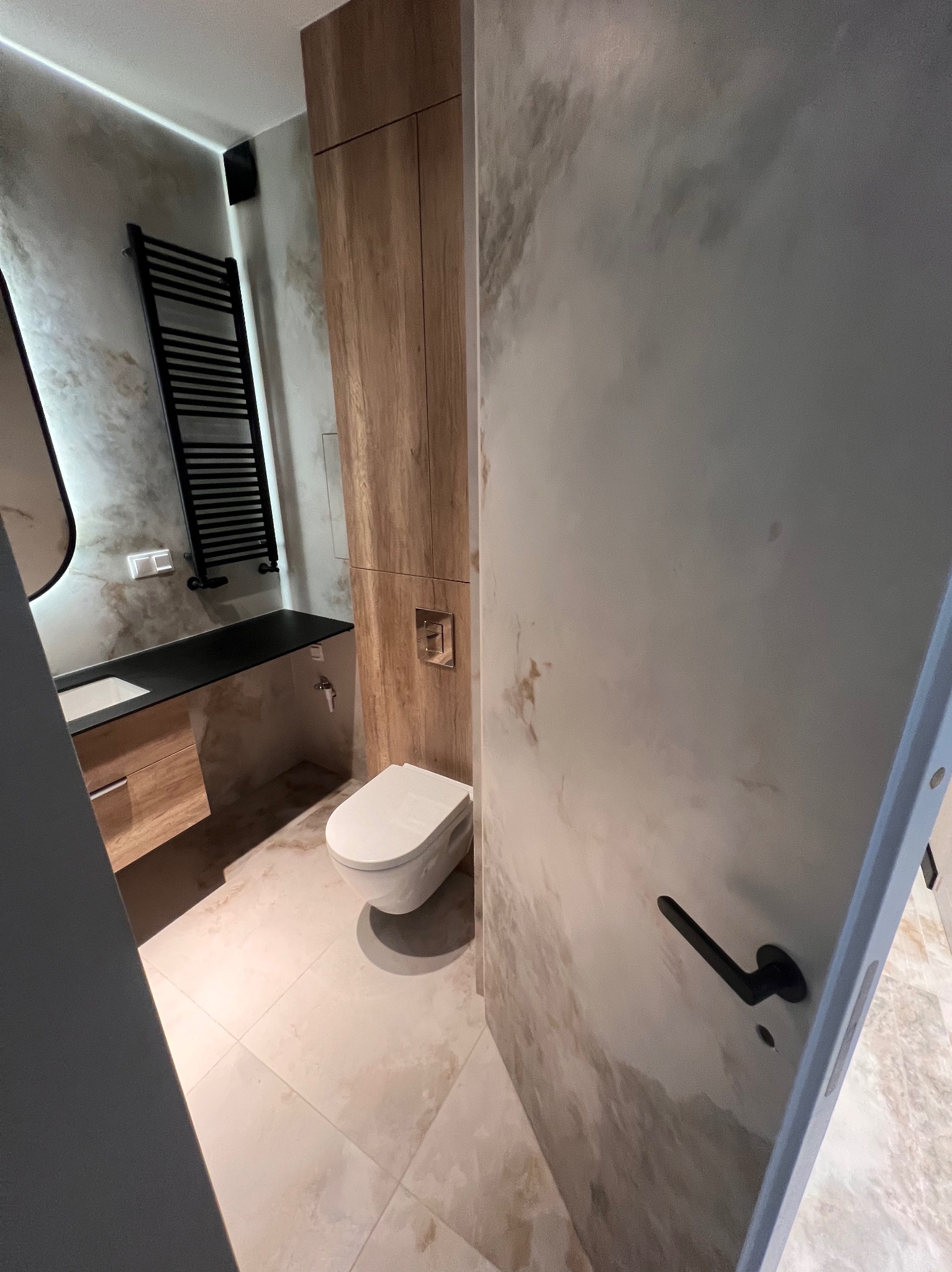 Elegancka łazienka z marmurkowymi ścianami, drewnianą zabudową i czarnym grzejnikiem drabinkowym. Umywalka z czarnym blatem i wisząca toaleta.
