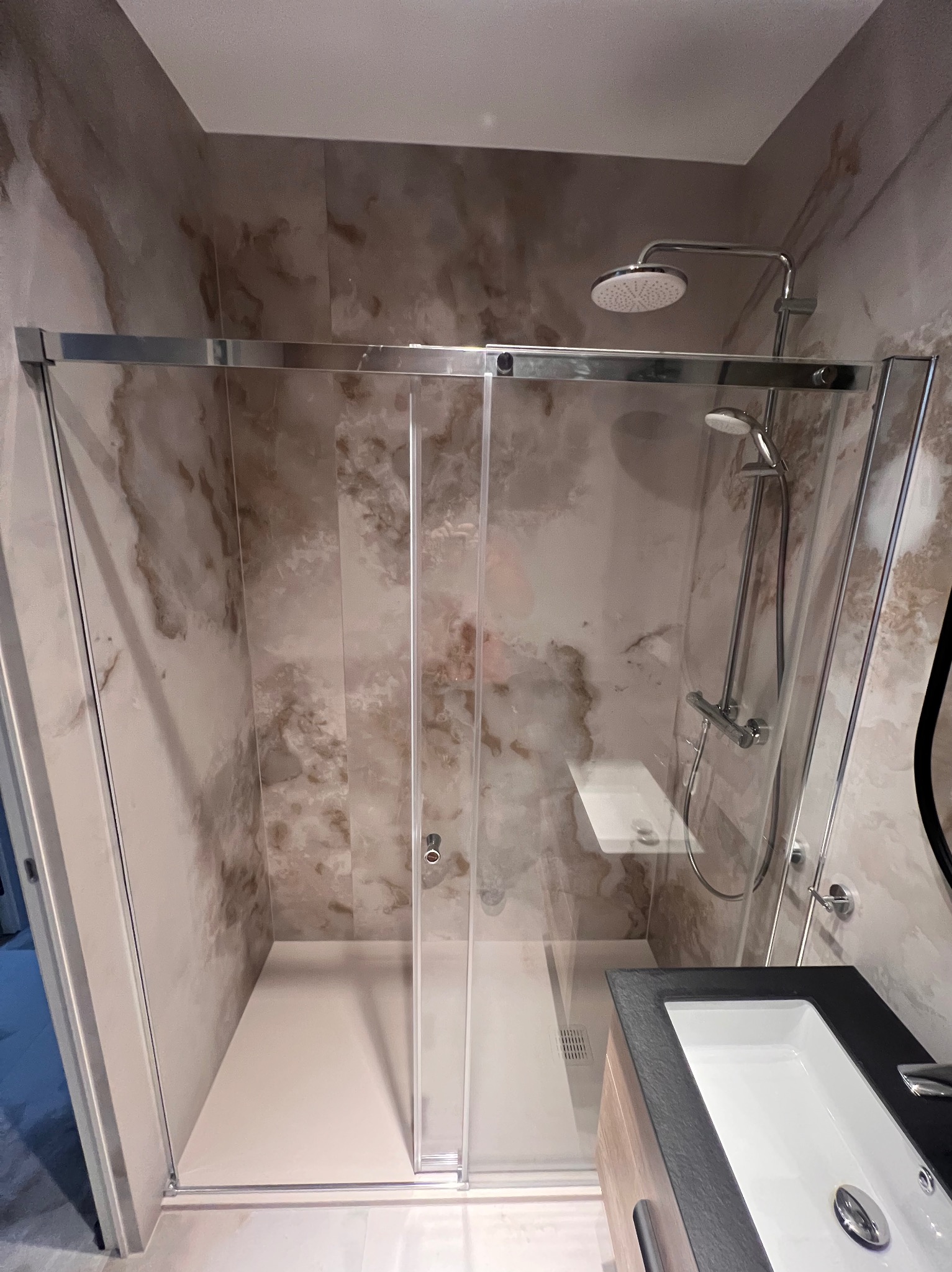 Nowoczesna kabina prysznicowa z przesuwnymi drzwiami i marmurowymi płytkami w beżowych odcieniach. Widoczna umywalka z czarnym blatem i chromowaną armaturą.