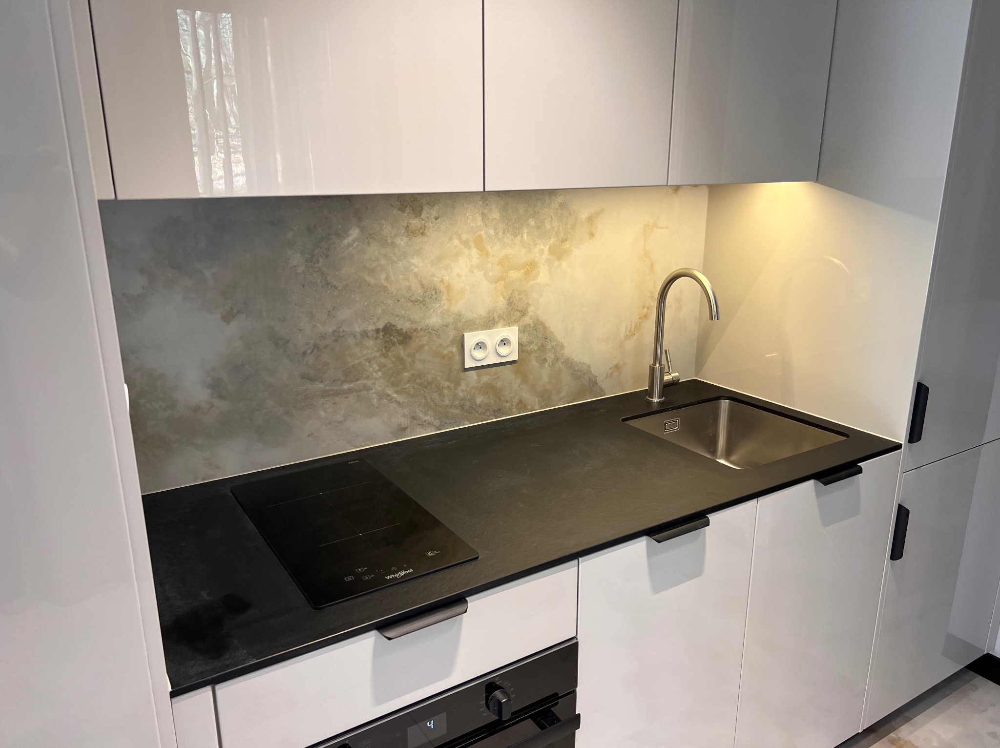 Minimalistyczna kuchnia z białymi szafkami, czarnym blatem i indukcyjną płytą. Na ścianie dekoracyjny panel imitujący marmur, oświetlenie punktowe pod szafkami.