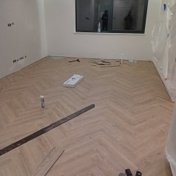 Jan Michurski Wykładziny Obiektowe - Układanie jasnych paneli w jodełkę francuską, widoczne narzędzia, listwy przypodłogowe jeszcze nie zamontowane, okno z odbiciami osób.