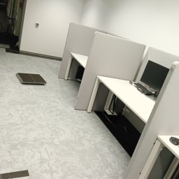 Jan Michurski Wykładziny Obiektowe - Układanie jasnoszarej wykładziny dywanowej w biurze z białymi biurkami i ściankami działowymi, widoczna niedokończona krawędź i stos płytek wykładzinowych.