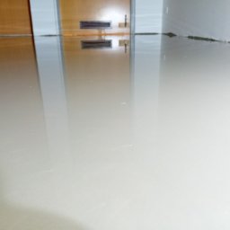 Jan Michurski Wykładziny Obiektowe - Wylewka samopoziomująca na podłodze, odbijająca drzwi i fragment ściany, przygotowana pod dalsze prace wykończeniowe.